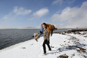 Man in a plaid shirt carries a laughing woman over his shoulder along a snowy lakeshore under a bright blue sky.