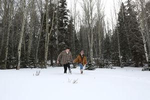 Couple holds hands and walks through deep snow in a peaceful winter forest of tall aspen and pine trees.
