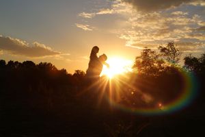 Silhouette of a person holding a child at sunset with golden sunbeams and a rainbow lens flare over a field.