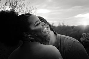 Black and white portrait of a smiling woman with hoop earrings embracing a man outdoors at dusk.