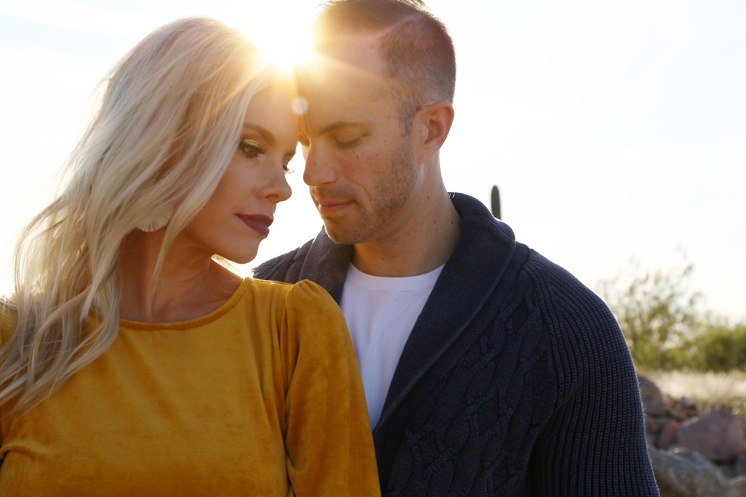 A blonde woman in a mustard top and a man in a navy cardigan stand close, foreheads touching, in warm golden sunlight.