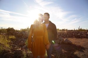 Couple embracing outdoors in a desert setting as bright sun flares behind them, the woman wearing a mustard yellow dress.