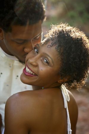 Smiling woman with short curly hair looks over her shoulder as a man kisses her cheek in warm outdoor light.