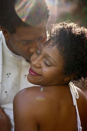 Woman with curly hair smiles contentedly with closed eyes as a man kisses her cheek in glowing sunlit light.