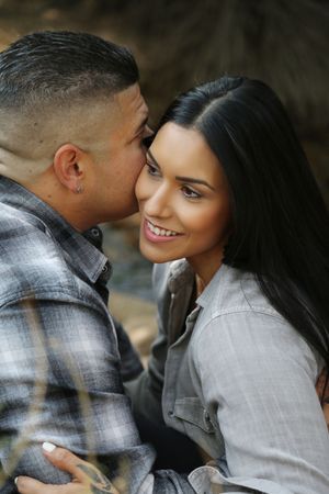 Smiling woman with long dark hair receives a kiss on the cheek from a tattooed man in a plaid flannel shirt.