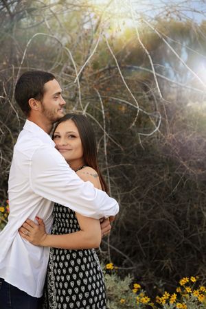 Engaged couple embracing among desert shrubs and yellow wildflowers, the man in a white shirt and woman in a patterned dress.