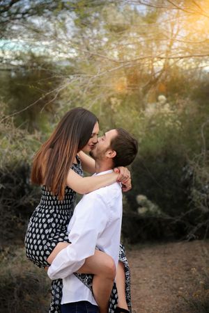 Couple kissing outdoors as a man lifts a woman in a polka dot dress, with warm golden light filtering through desert trees.
