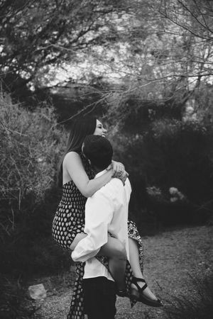 Black and white photo of a man lifting a woman in a polka dot dress as she wraps her arms around him on a wooded path.