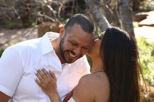 Smiling bearded man in a white shirt laughs as a dark-haired woman kisses his cheek during an outdoor couple photoshoot.