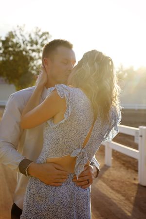 Couple in a tender embrace shares a kiss outdoors as golden sunlight glows behind them, the woman wearing a blue floral dress.