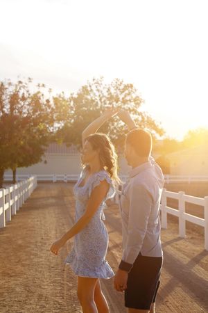 A couple dances and twirls on a sunlit dirt path lined with white fences as golden evening light glows behind them.