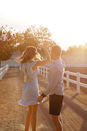 A woman in a floral dress twirls under her partner's raised arm on a sun-drenched ranch path at golden hour.