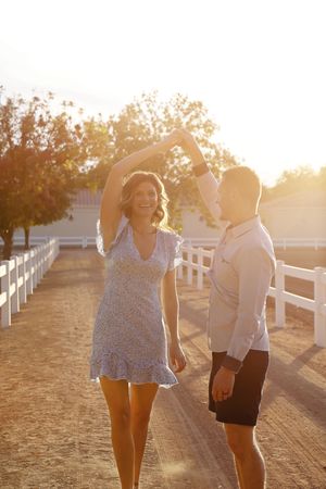 Smiling woman in a blue floral dress twirls under a man's raised arm on a sunlit dirt path beside a white ranch fence.