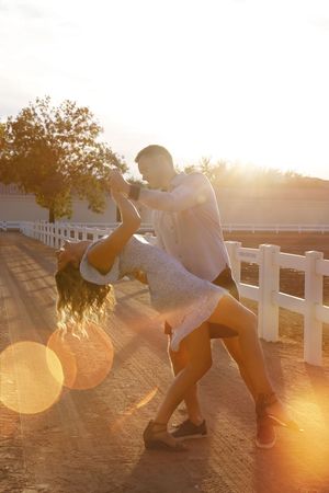 Man in a light button-up shirt dips a woman in a floral dress during a romantic dance at golden hour near a white fence.