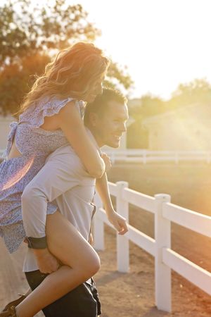 Laughing man gives a woman in a floral dress a piggyback ride along a white fence in warm golden sunset light.