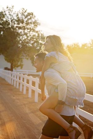 Woman in a floral dress rides piggyback on a smiling man along a dirt path lined with a white fence at sunset.