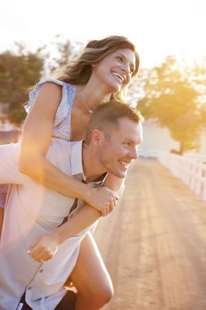 Joyful couple laughs together as a smiling woman gets a piggyback ride outdoors in warm golden sunlight.