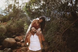 Woman in a rust top sits on the shoulders of a smiling man in a white shirt amid boulders and brush on a hiking trail.