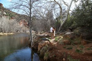 Couple embraces on a rocky creek bank beside calm water, surrounded by bare trees and red rock cliffs in the distance.