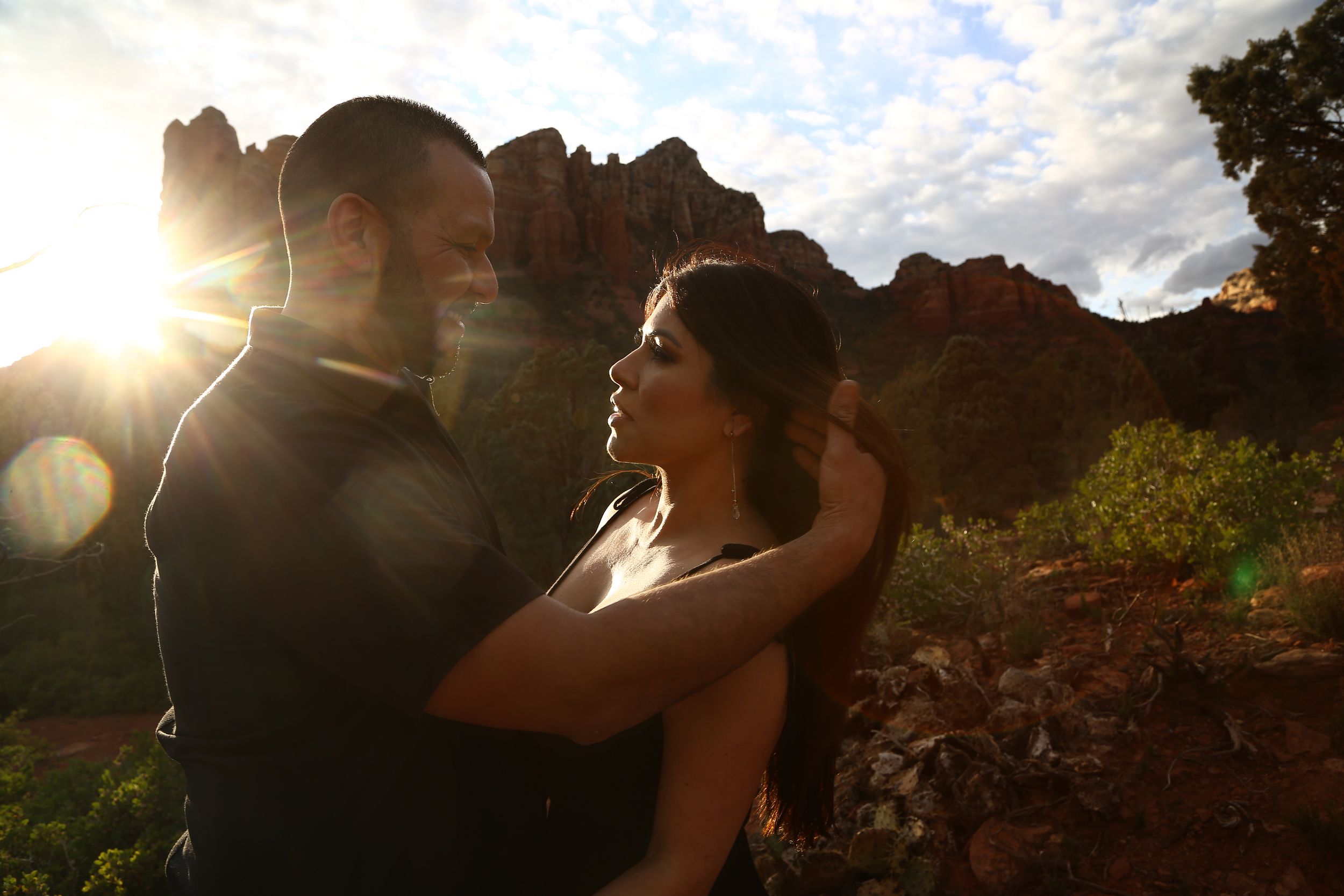 Couple in a tender embrace is backlit by a glowing sunset with red rock buttes and scattered clouds behind them.