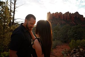 Bearded man in a black shirt holds a long-haired woman close at sunset with red rock formations and pine trees behind.