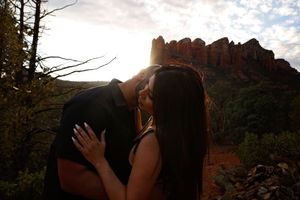 Couple kissing at sunset in front of red rock formations in a desert landscape with trees in the foreground.