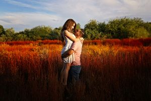 Man lifts a woman in a white dress as they embrace in a field of glowing red grass under a blue sky.