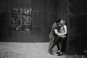 Black and white photo of a couple kissing beside a weathered dark wall with a boarded wooden window.