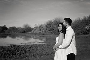 A black and white engagement photo of a couple embracing beside a calm pond with shrubs and trees in the background.