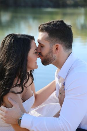 A bearded man in a white shirt kisses the forehead of a smiling dark-haired woman in a white dress beside a lake.