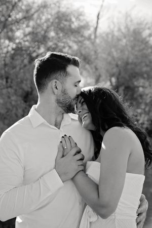 A black and white portrait of a laughing woman with an engagement ring as her fiance kisses her forehead outdoors.