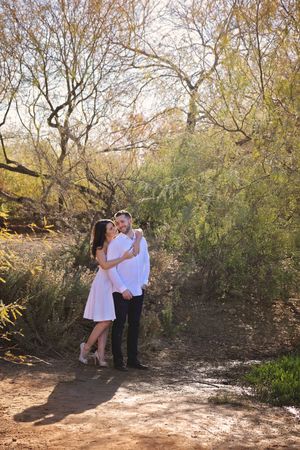 An engaged couple in white outfits embraces on a dirt path surrounded by desert trees and golden-lit shrubs.