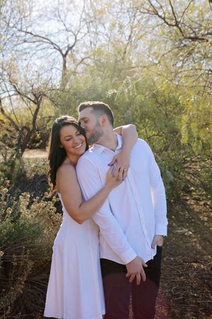 A smiling woman in a white dress hugs a bearded man in a white shirt among desert trees and shrubs in soft sunlight.