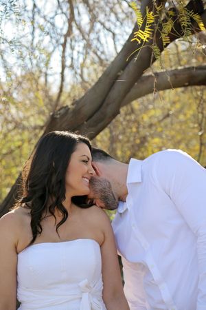 A woman in a white strapless dress smiles as her fiance in a white shirt nuzzles her cheek beneath a tree branch.