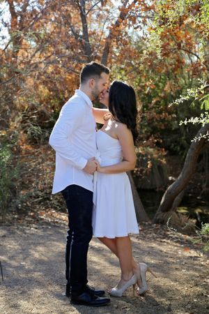 Couple in white outfits share a romantic kiss on a dirt path surrounded by colorful autumn trees during an engagement photo session.