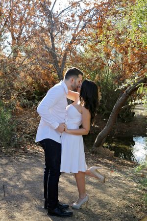 Woman in a white dress lifts her heel while kissing a man in a white shirt beside a pond and fall foliage in an outdoor engagement shoot.