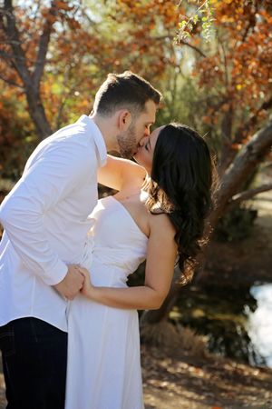 Close-up of an engaged couple kissing near a calm pond, with the woman in a white dress and the man in a white button-up shirt.