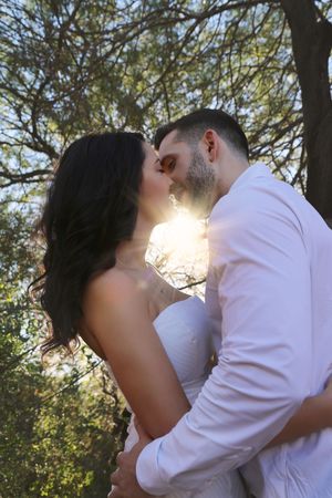 Romantic couple in white about to kiss outdoors under tall trees as golden sunlight glows through the branches.