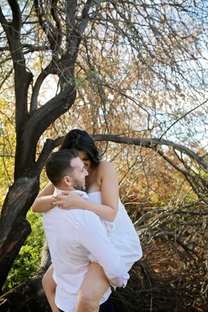 Man in a white shirt lifts a woman in a white dress in his arms beneath a twisted tree during a romantic outdoor couple photo shoot.
