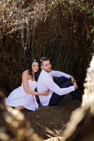 Smiling couple in white outfits sitting together on the ground beneath tall dry grasses in an outdoor engagement photo.
