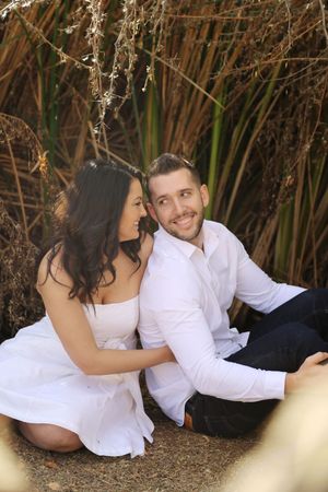 Smiling couple in white outfits sit close together on the ground in front of tall reeds, sharing a happy moment in an engagement photo.