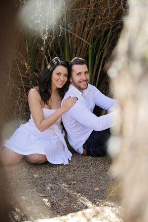 Happy engaged couple in white sit together on the ground and smile at the camera, framed by tall reeds and soft blurred foreground branches.