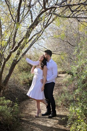 Couple embracing on a sunlit dirt trail beneath arching trees, the woman in a white dress reaching up toward the branches.