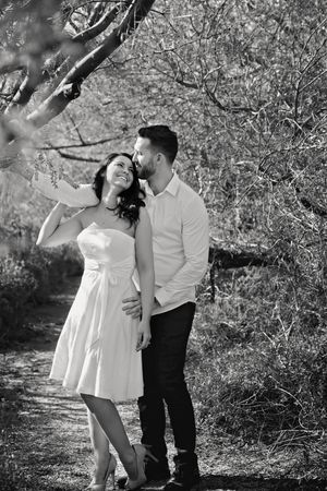 Black and white photo of a smiling woman in a white dress embraced by a man in a white shirt on a wooded path during an engagement session.
