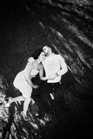 Black and white overhead photo of an engaged couple lying close together on the ground as he kisses her forehead.