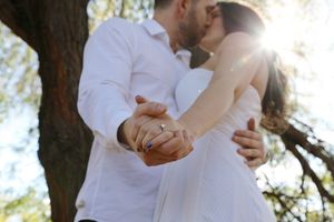 Close-up of a couple in white embracing in warm sunlight, their clasped hands showing a ring beside a large tree.