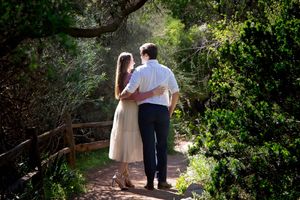 Couple in white clothing walking arm in arm along a shaded dirt path surrounded by lush green trees and wooden fencing.