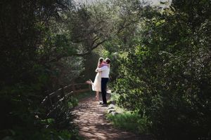Couple embracing and kissing on a wooded trail framed by dense green trees and dappled sunlight.