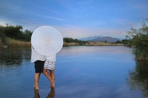 Couple stands ankle-deep in a calm lake kissing behind a white parasol, with distant mountains and blue sky.