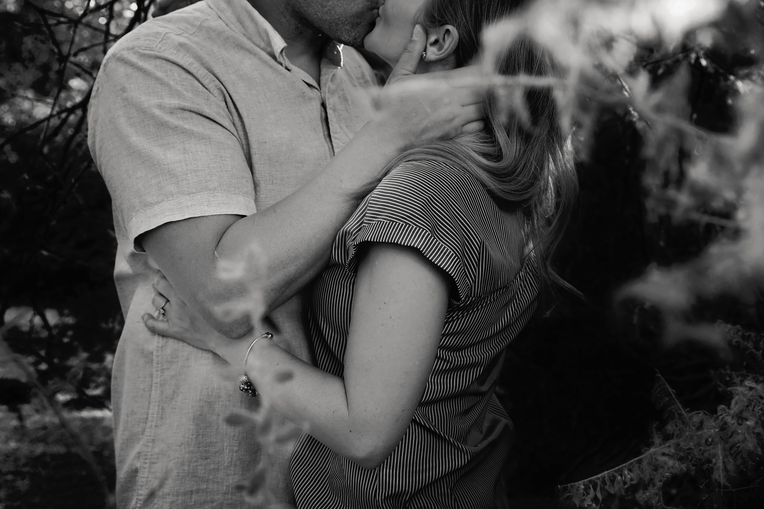 Black and white close-up of a couple kissing and embracing, framed by blurred foliage in the foreground.